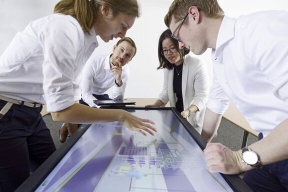 There are a total of four people in the photo, two women and two men. They are bending over a mo-nitor with a technical model.