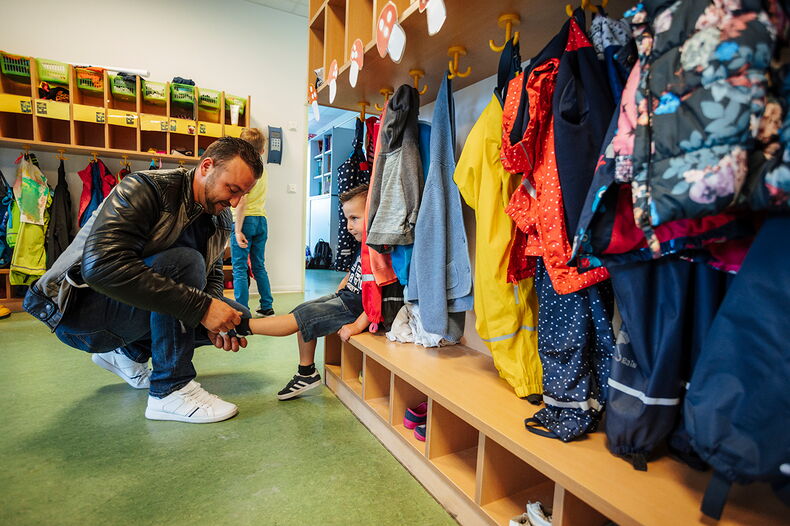 Vater zieht Kind an der Garderobe in der Kita die Schuhe an