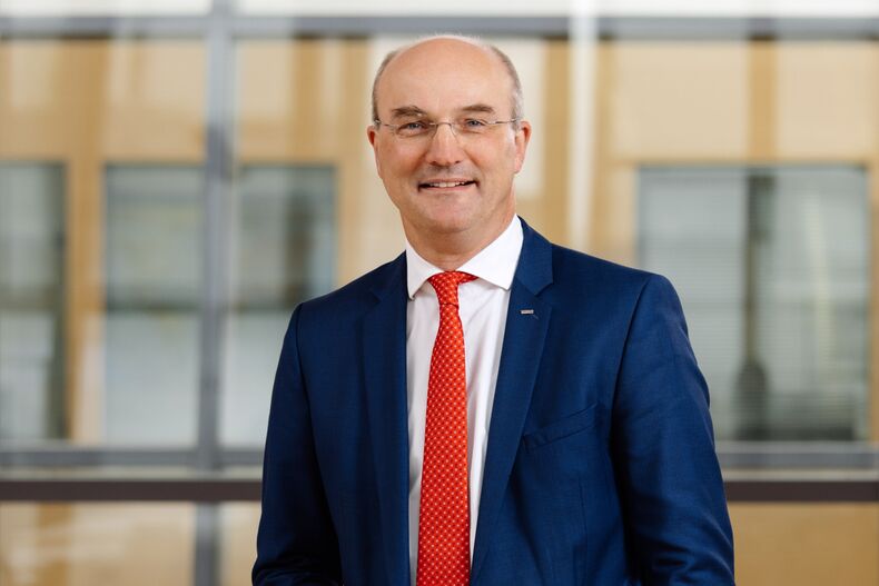 Professor Ulrich Rüdiger, Rector of RWTH, stands in front of a glass wall. He is wearing a dark blue suit, a white shirt and a red tie. He smiles into the camera.