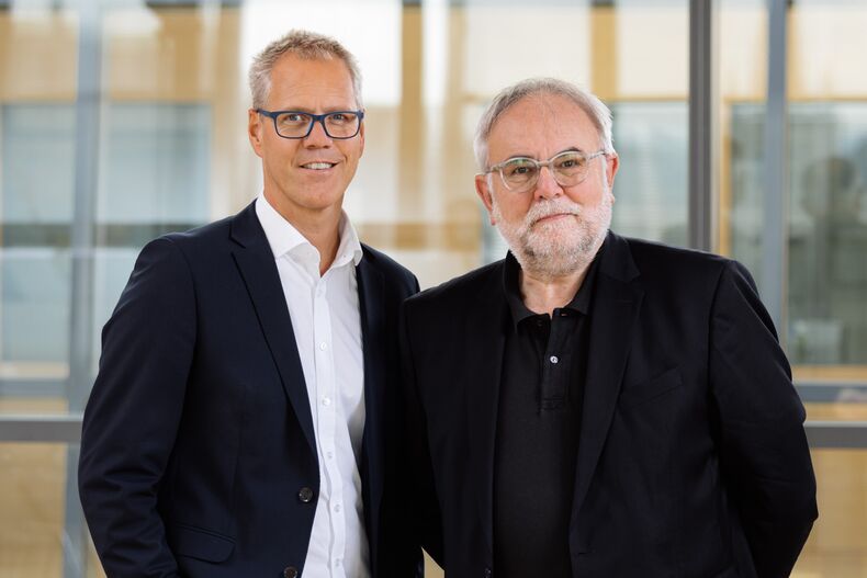 Photo of permanent chancellor representative Thomas Trännapp and chancellor Manfred Nette-koven in front of a glass wall. Thomas Trännapp with black jacket, white shirt and jeans. Manfred Nettekoven with dark blue jacket, black polo shirt and jeans. Both are smiling at the camera.