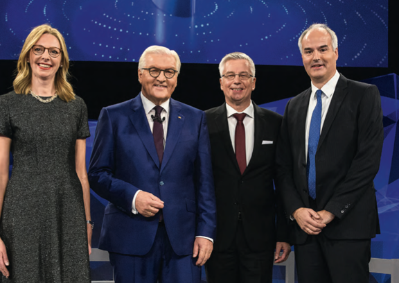 Drei Personen in Anzug und eine Person in Kleid. Alle stehen nebeneinander.