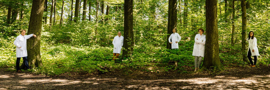 Zwei weibliche und drei männliche Personen in weißen Laborkitteln stehen in unterschiedlichen Posen an Bäume gelehnt verteilt im Wald. Sie lächeln in die Kamera.