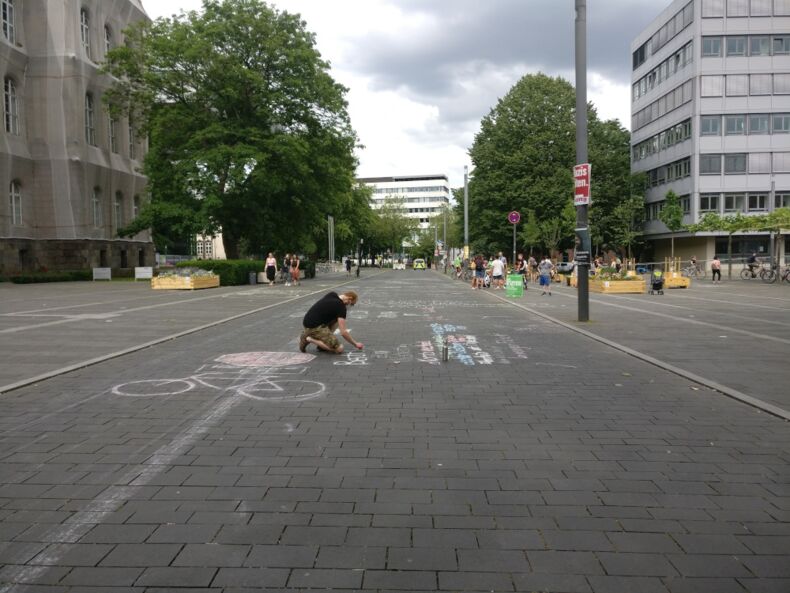 Das Bild zeigt eine Person, die auf dem Templergraben kniet und mit Kreide die Straße bemalt. Im Hintergrund sind Sitzgelegenheiten aus Holz auf dem Platanenplatz zu sehen.