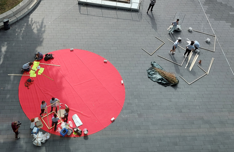 Das Bild zeigt eine Aufnahme aus der Vogelperspektive. Auf einem großen, runden roten Teppich bauen Teilnehmende der Summer School 2022 Kunstinstallationen auf und werden dabei gefilmt.