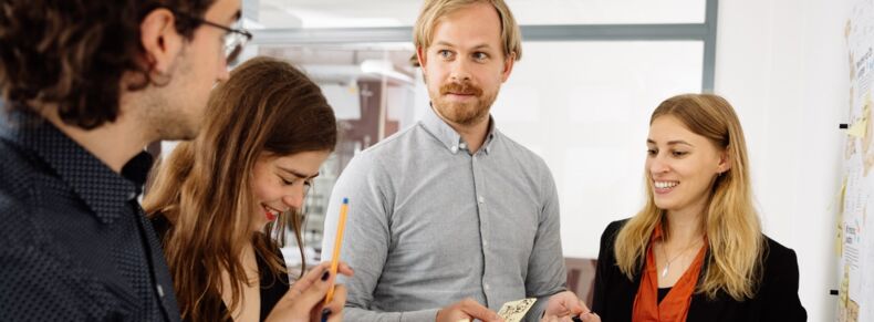 Das Bild zeigt vier Personen im Gespräch. Die Personen lächeln. Eine Person hält einen Notizzettel in der Hand.