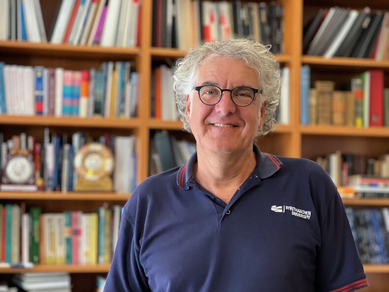 The picture shows Professor Dr. Reicherter. He is wearing a blue shirt with the inscription RWTH Aachen University. A bookshelf can be seen in the background.