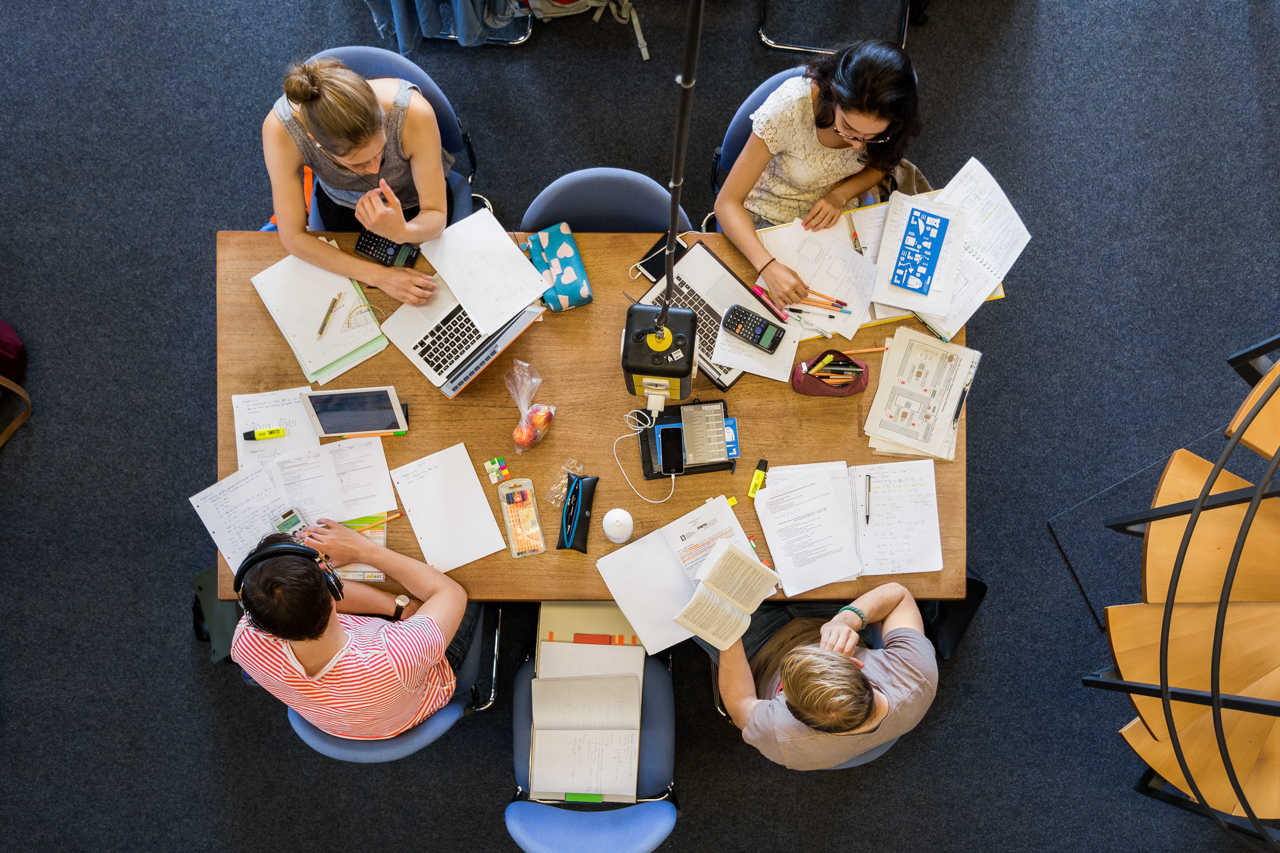 Zwei weibliche und zwei männliche Personen sitzen an einem Tisch. Sicht von oben. Auf dem Tisch sind Lernmaterialien ausgebreitet.